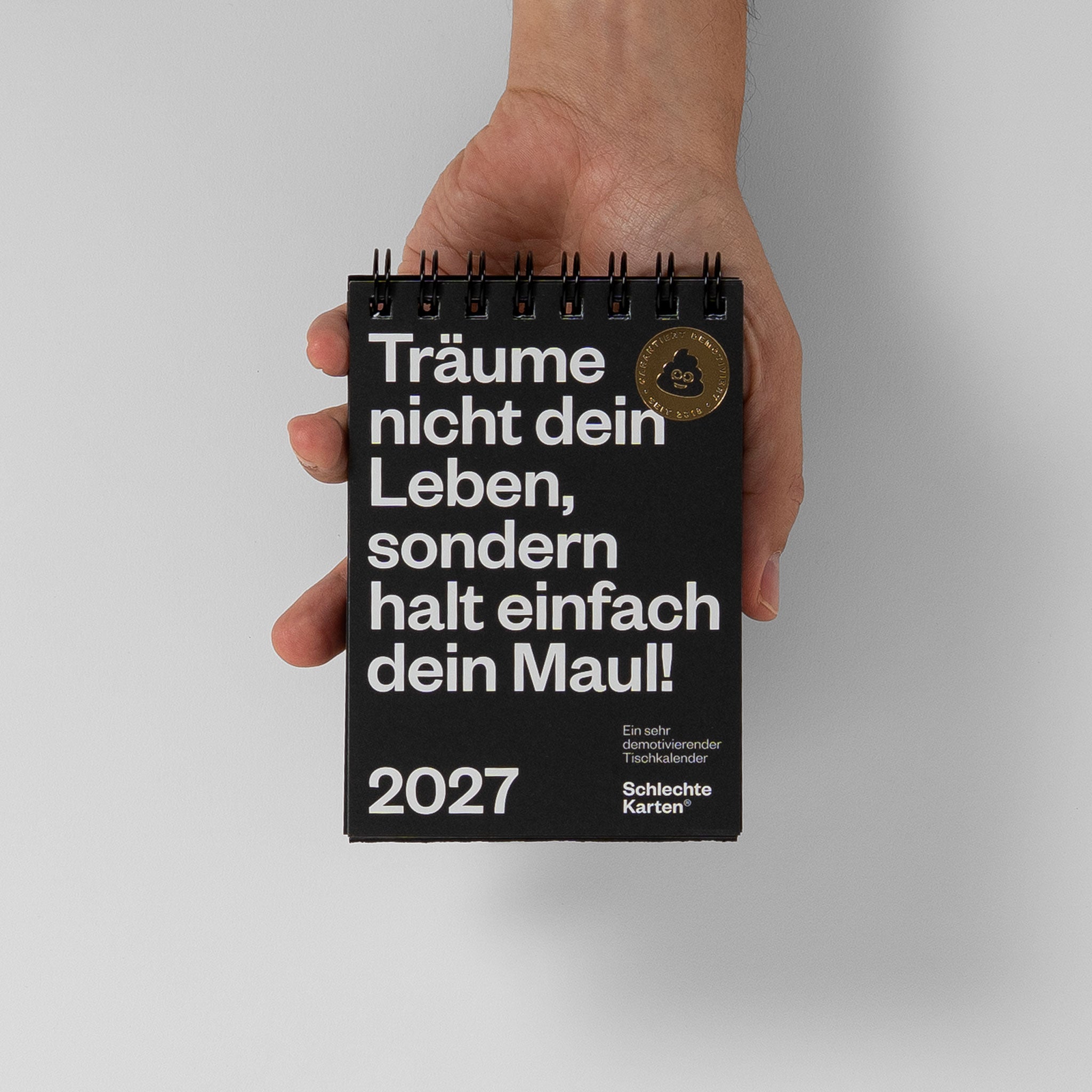 Größenvergleich des demotivierenden Tischkalenders 2027 mit einer Hand zum Maßstab